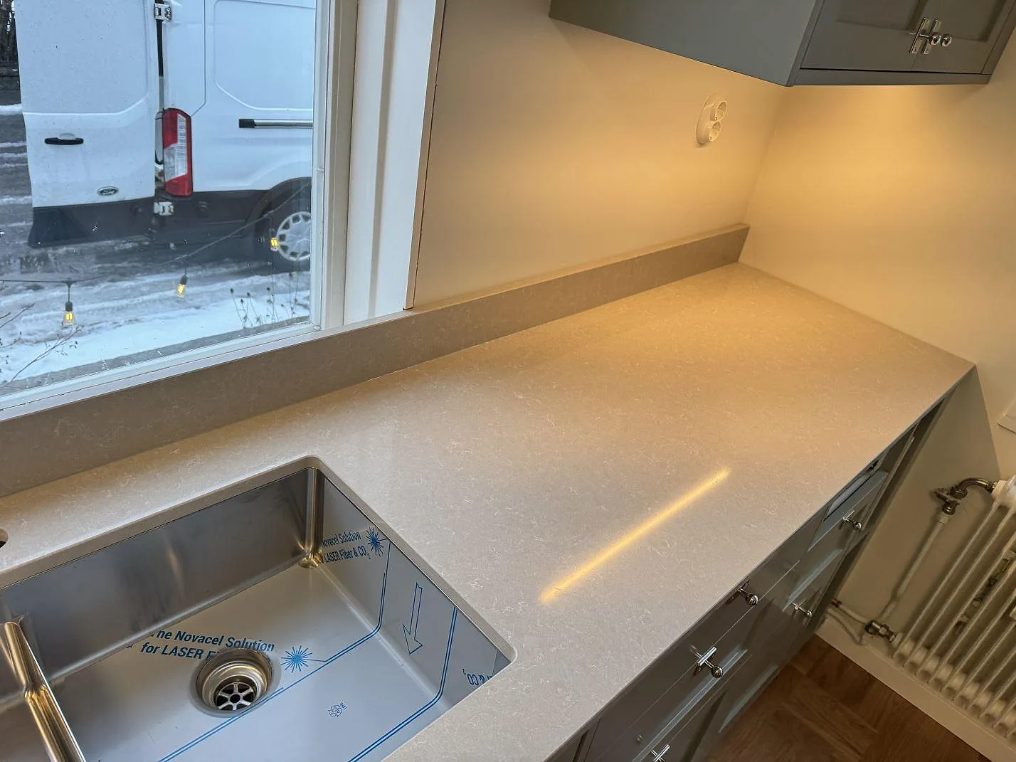 Köksbänkskiva i vit Technistone Noble Ivory White i Kalmar, eklik vy med fönster, underlimmad rostfri diskho och gråa shakerluckor under bänken.