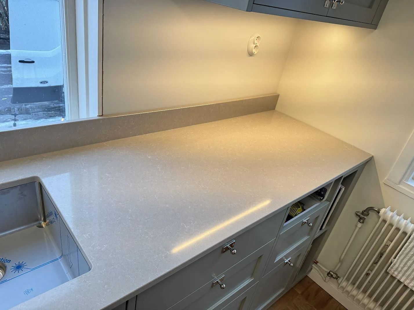 Köksö och bänkskiva i Technistone Noble Ivory White i ett kök i Kalmar, polerad vit yta med rostfri dubbelho vid fönstret och radiator i hörnet.