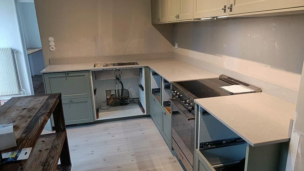 Köksbänkskiva i ljus Technistone Noble Ivory White i ett L-format kök i Klagshamn, fotograferad under monteringen med salviegröna stommar och fristående induktionsspis.