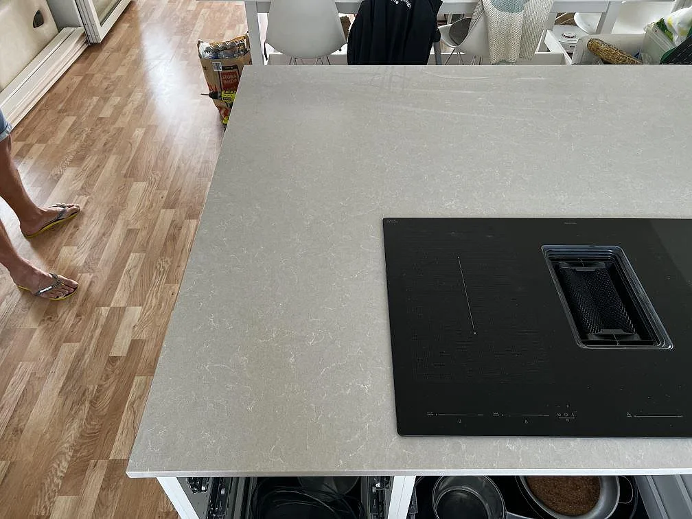 Installerad köksö i Technistone Noble Ivory White, polerad komposit, med svart häll och öppna underskåp i Klagshamn.