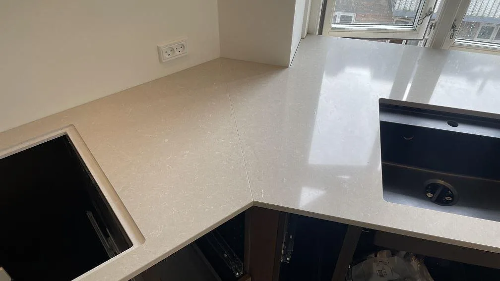 L-formad köksbänkskiva i polerad Technistone Noble Ivory White komposit med hörnskarv, svart diskho och hälluttag vid fönster i Köpenhamn.