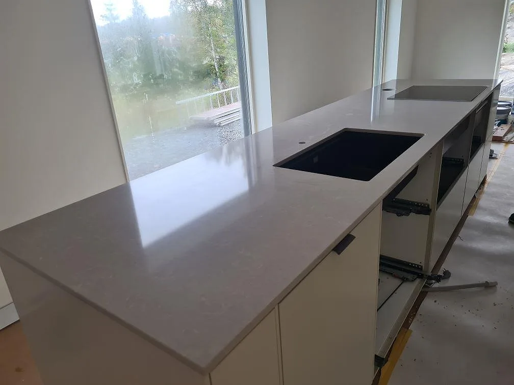 Köksbänkskiva i Technistone Noble Ivory White, polerad kvartskomposit, längs stora fönster med svart underlimmad diskho och induktionshäll i Ösmo.