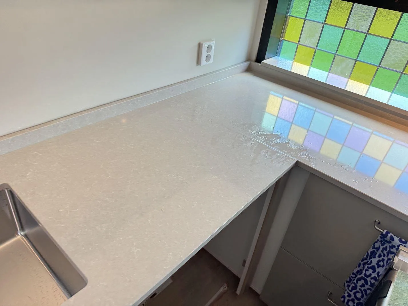 L-formad köksbänkskiva i polerad Technistone Noble Ivory White med ljus ådring och färgat fönsterglas i Oskarström.