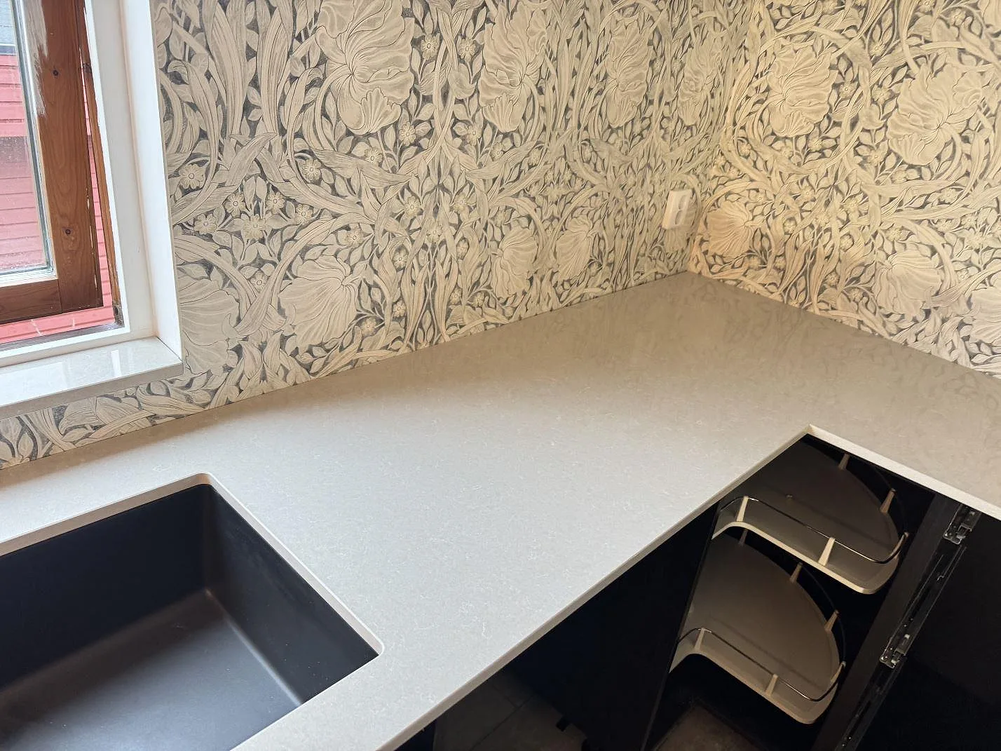 L-formad köksbänkskiva i Technistone Noble Ivory White, polerad kvartskomposit, med mörk diskho och fönsterbänk i samma sten i Trelleborg.