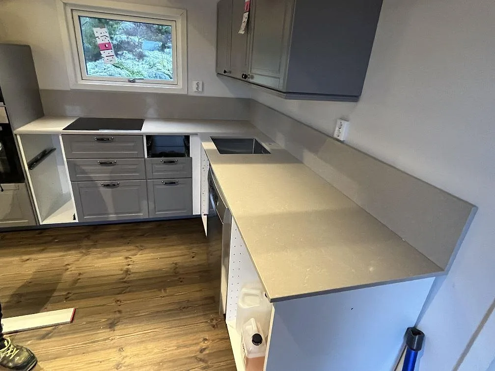 Köksbänkskiva i 20 mm polerad Technistone Noble Ivory White, ljus komposit, med häll, diskho och låg stänkkant i ett U-format kök i Vikbolandet.
