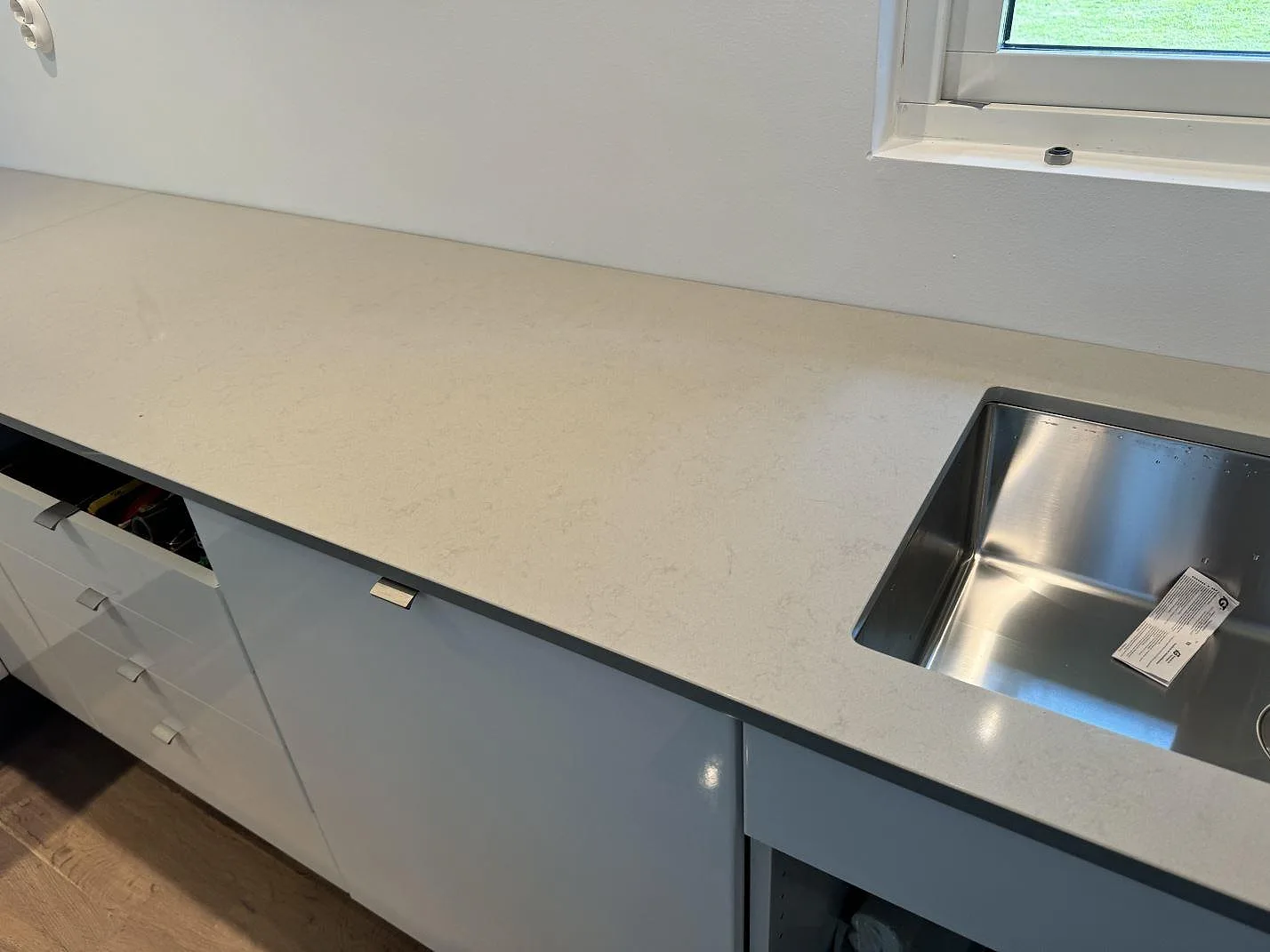 Lång köksbänkskiva i polerad Technistone Noble Portland Grey komposit med rostfri diskho vid fönster i Räng.