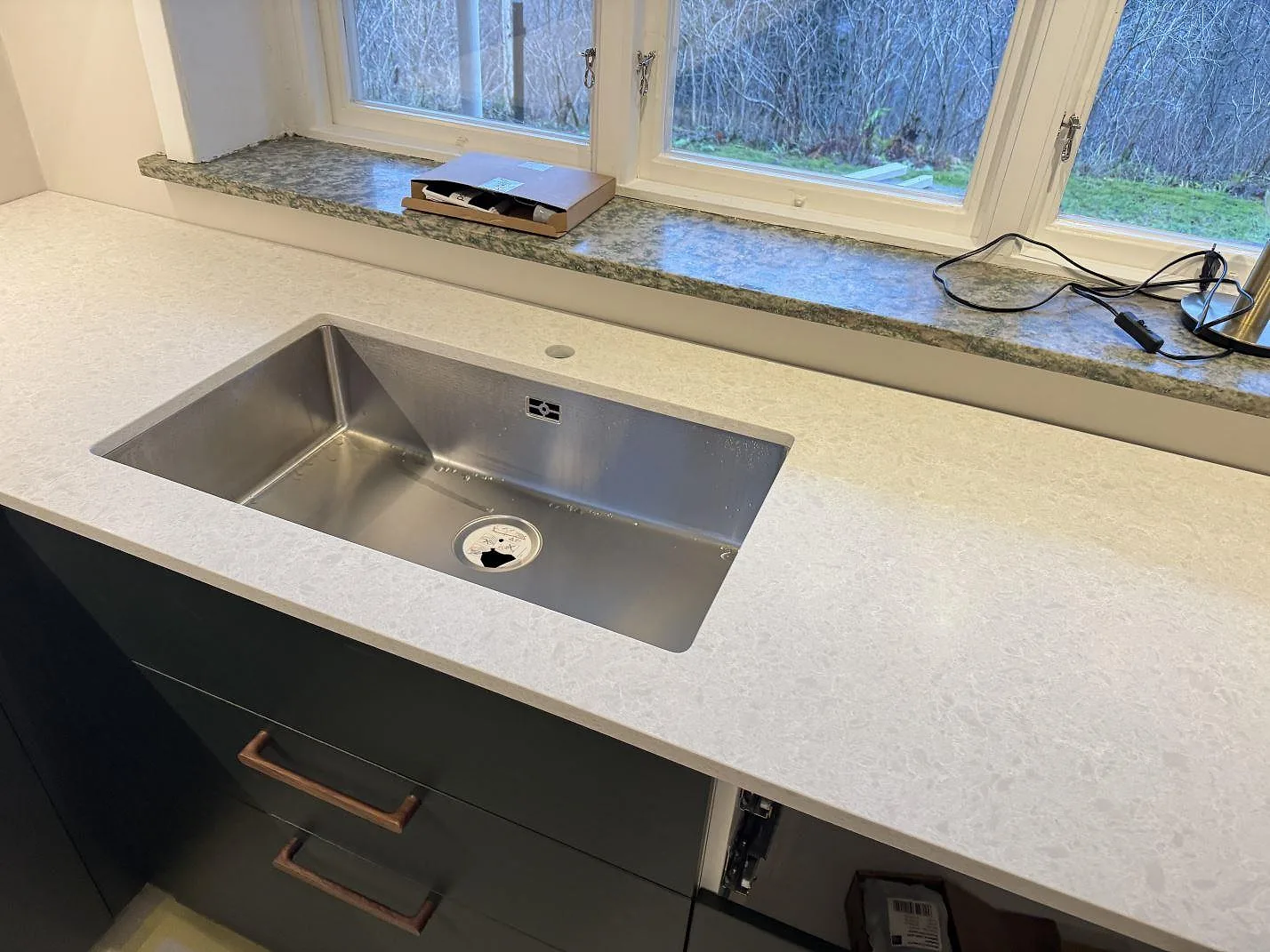 Köksbänkskiva i Technistone Noble Quartzite, matt vitgrå kvartskomposit, med underlimmad rostfri diskho vid fönstret i ett kök i Norrahammar.