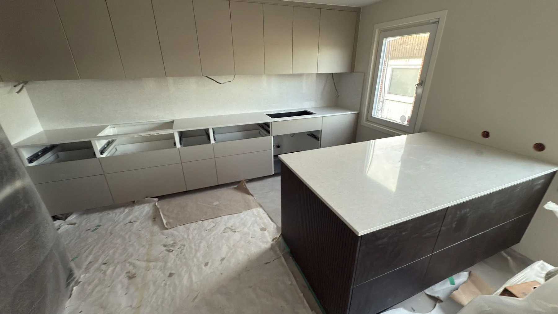 Köksbänkskiva i ljusgrå Technistone Noble Quartzite, polerad kvartskomposit, med stor köksö och uppvikt stänkkant i ett kök under montering i Södertälje.