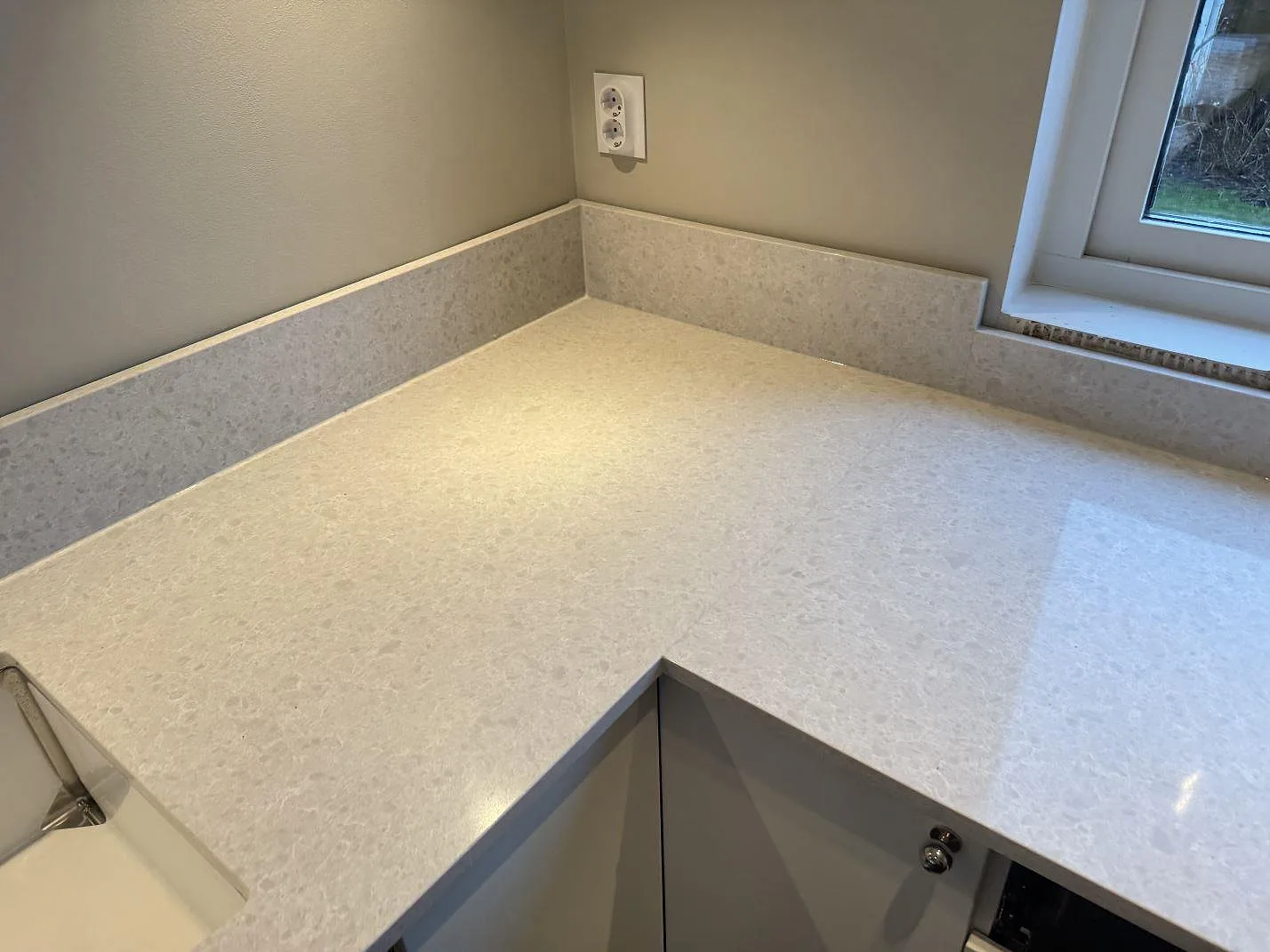 L-formad bänkskiva i polerad Technistone Noble Quartzite komposit med kort stänkskydd vid fönster i Västra Frölunda.