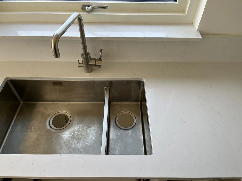 Underlimmad rostfri dubbeldiskho med borstad köksblandare i vit Technistone Noble Supreme White bänkskiva vid fönstret i ett kök i Beddingestrand.