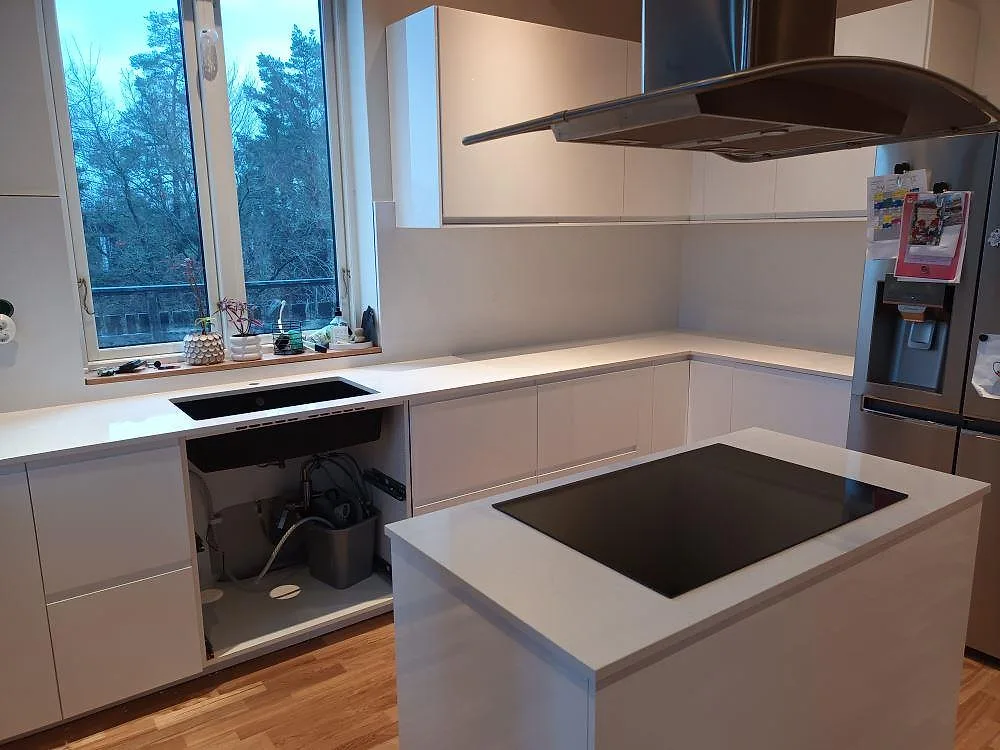 Vit polerad köksbänkskiva i Technistone Noble Supreme White med underlimmad svart diskho under fönstret och köksö med induktionshäll i Huddinge.