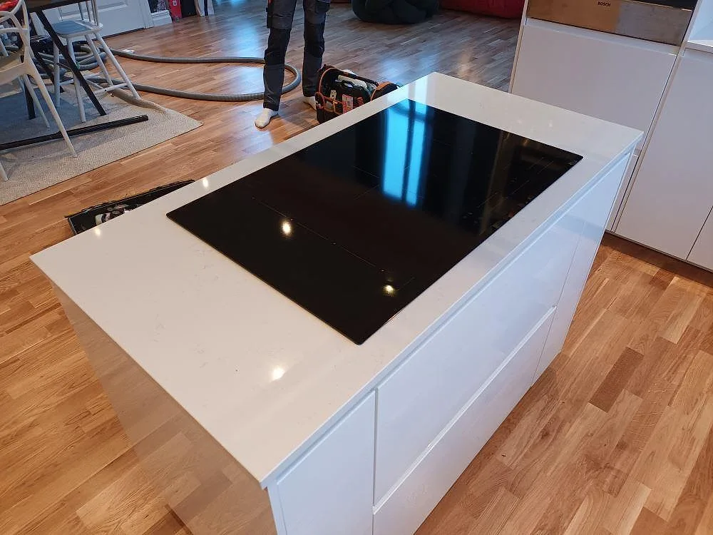 Köksö i vit Technistone Noble Supreme White med infälld svart induktionshäll, sedd ovanifrån under montering i ett kök i Huddinge.