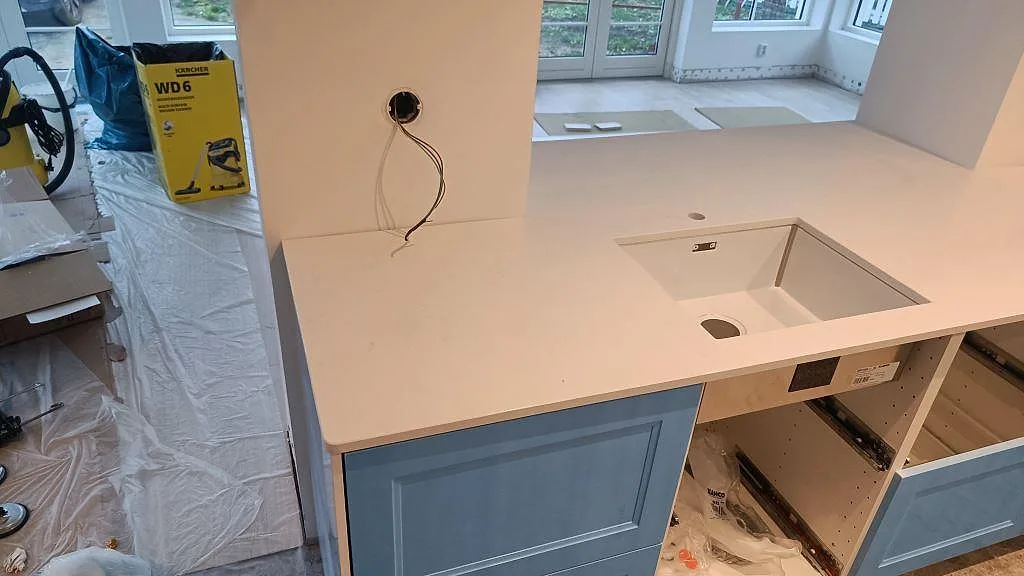 Köksbänkskiva i vit Technistone Noble Supreme White med underlimmad diskho och uppvikt stänkkant i ett kök under montering i Tygelsjö.