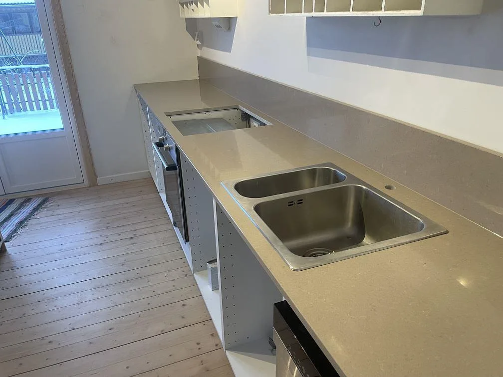 Polerad köksbänkskiva i greige Technistone Noble Troya med överlimmad rostfri dubbelho och uppvikt stänkkant i ett kök i Karlstad.