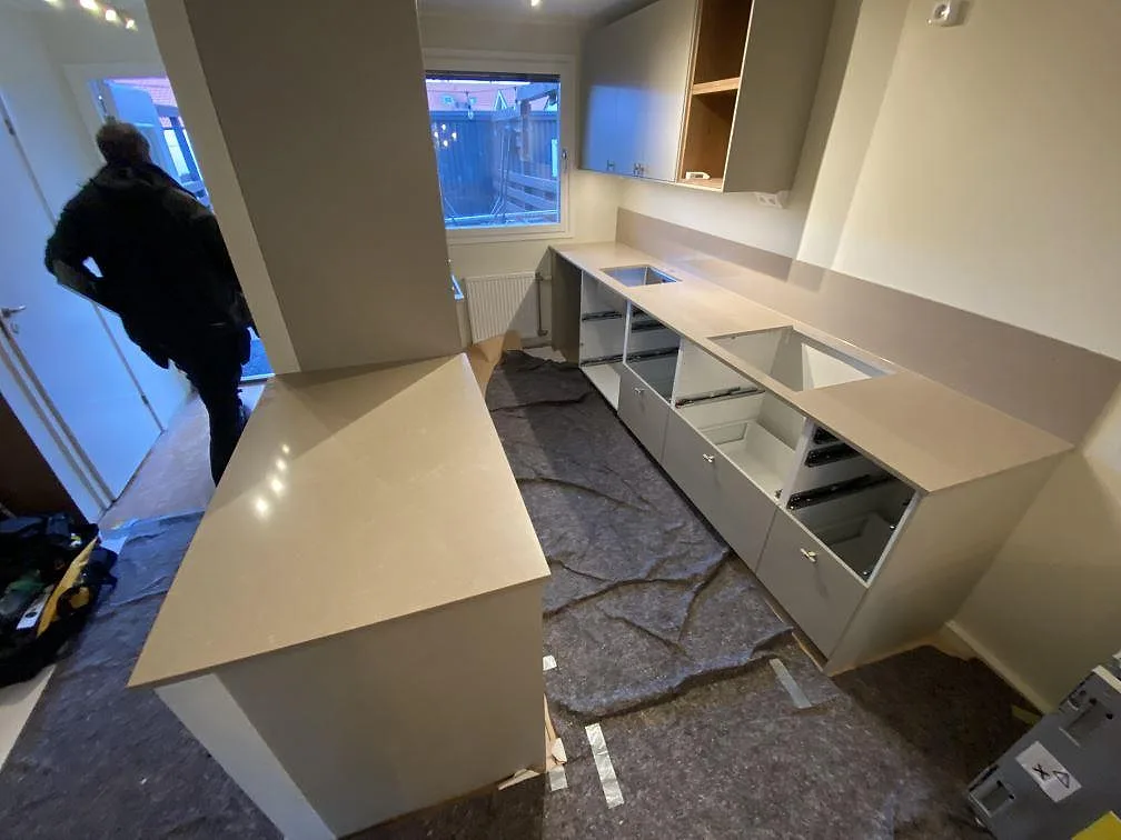 Köksbänkskiva i greige Technistone Noble Troya, polerad komposit, i ett L-format kök i Tullinge med halvö, underlimmad diskho och plats för induktionshäll under montering.