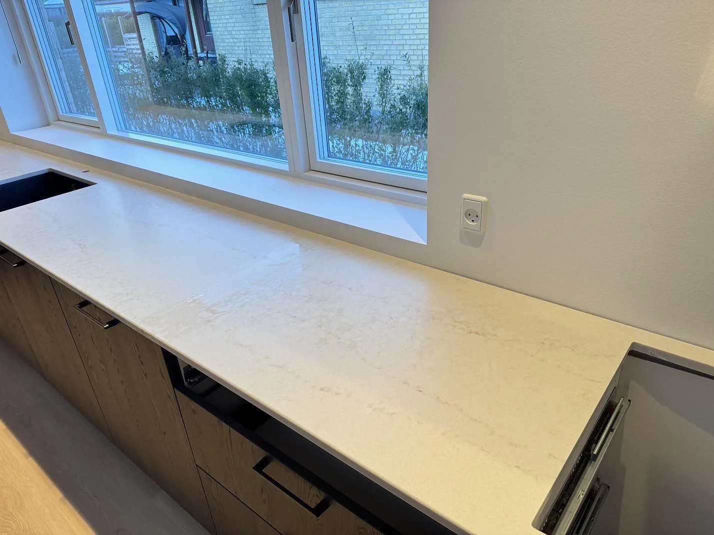 Lång köksbänkskiva i marmorlik Technistone Noble Vintage längs fönsterbandet med mörka ekluckor i ett kök i Glostrup.