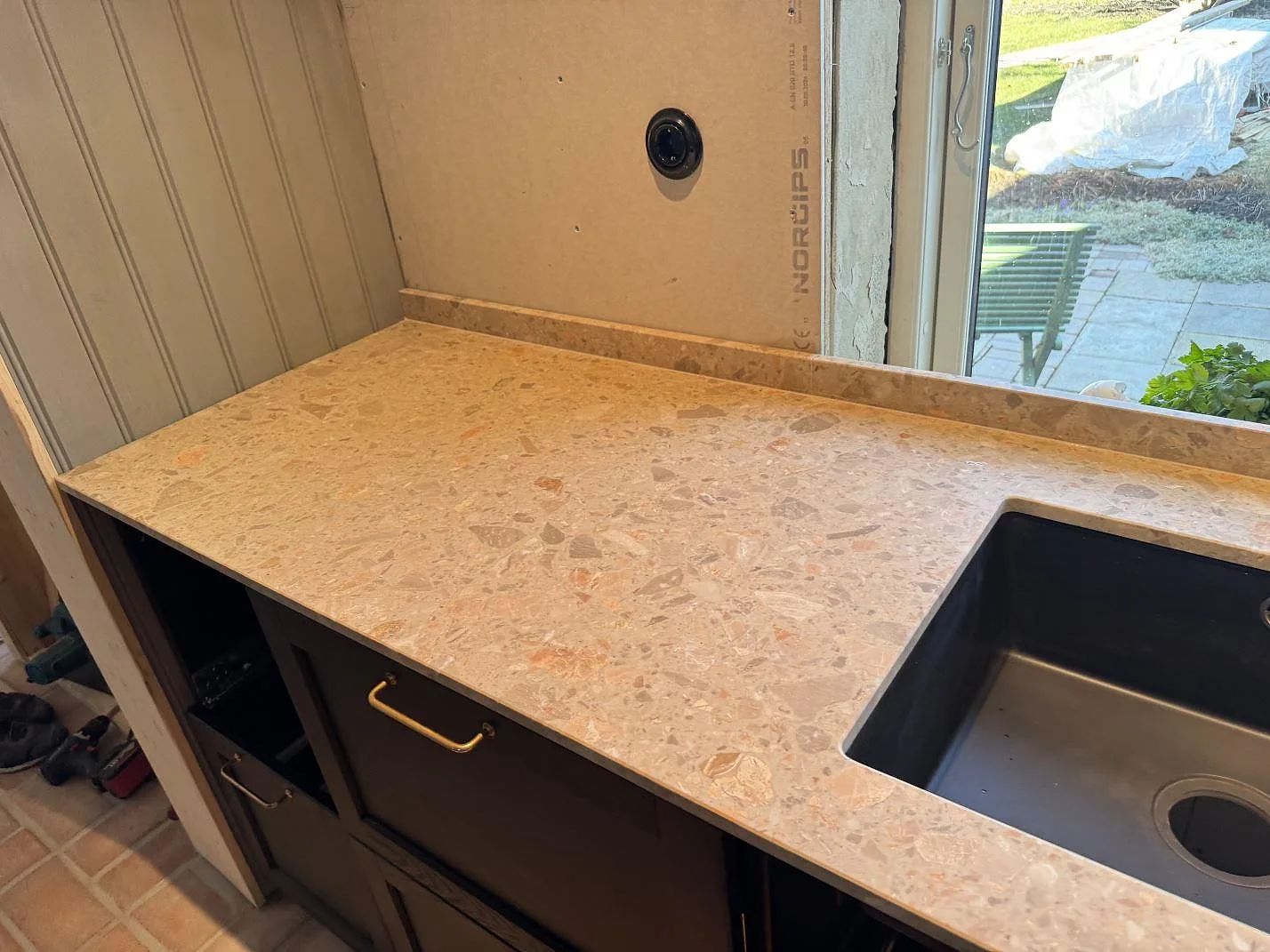 Matt köksbänkskiva i Terrazzo Breccia Oniciata med svart diskho och uppvikt stänkkant vid fönster i Tomelilla.