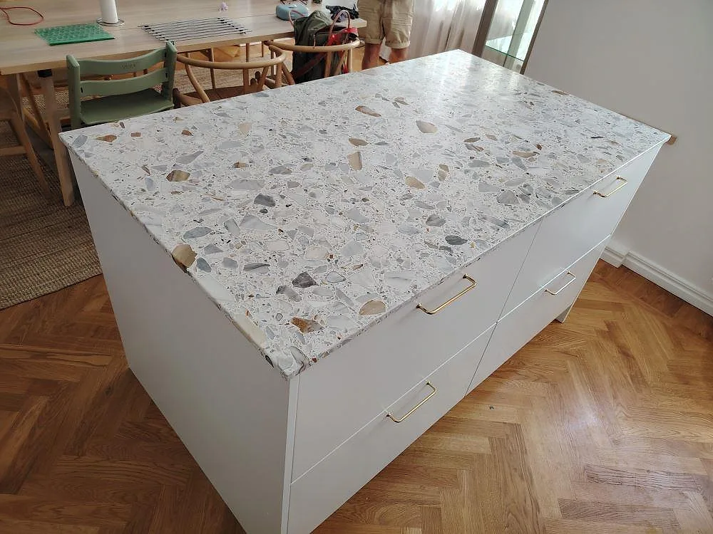 Närbild på matt köksbänkskiva i Terrazzo Calacatta på köksö, med färgade marmorfragment, vita lådfronter och matplats bakom i Sundbyberg.