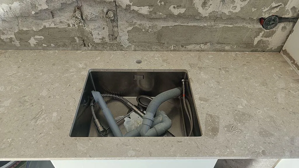 Underlimmad rostfri diskho i en matt köksbänkskiva av Terrazzo Perlato Royal mot ofärdig betongvägg i Ystad.