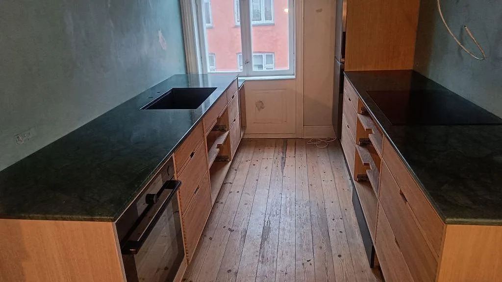 Köksbänkskiva i Verde Guatemala, polerad grön marmor, i ett galleykök under montering i Frederiksberg med två bänkrader och stort fönster.