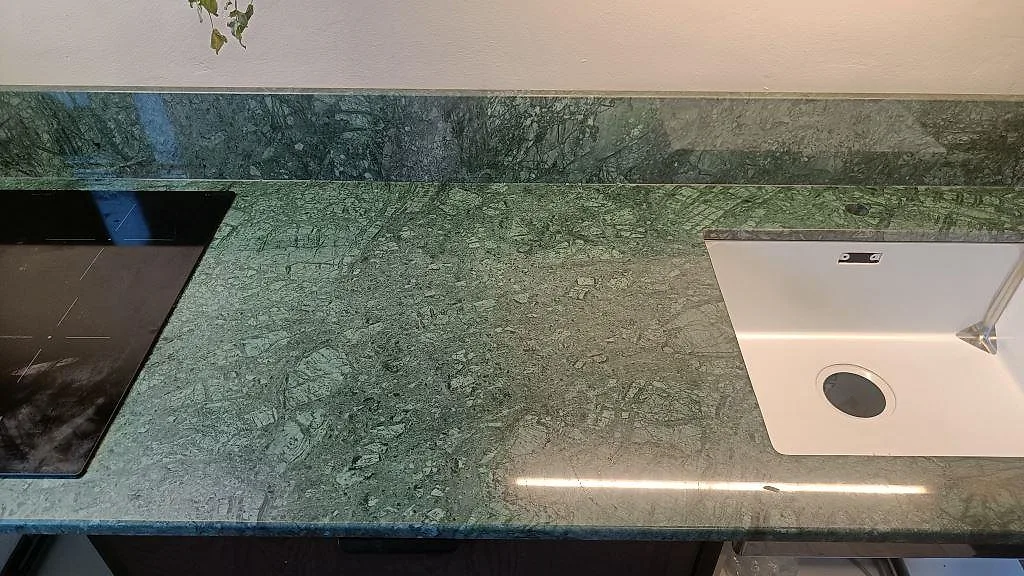 Närbild uppifrån av polerad Verde Guatemala marmorbänkskiva med synlig ådring, induktionshäll, vit diskho och stänkkant i Malmö.