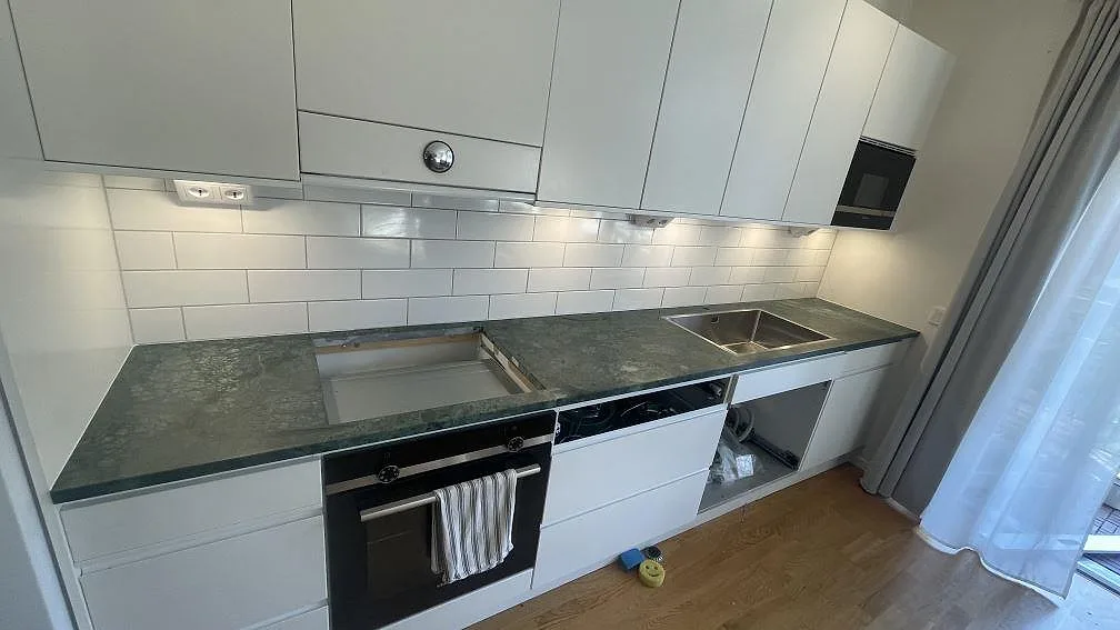 Köksbänkskiva i grön Verde Guatemala marmor, matt yta, längs vita köksluckor med vitt tegelfogat stänkskydd, rostfri underlimmad diskho och urtag för häll i ett kök i Malmö.