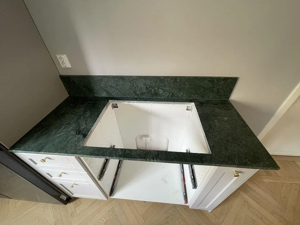 Detalj av matt köksbänkskiva i Verde Guatemala marmor med tydlig grön ådring, öppet skåp och stänkkant i Uppsala.