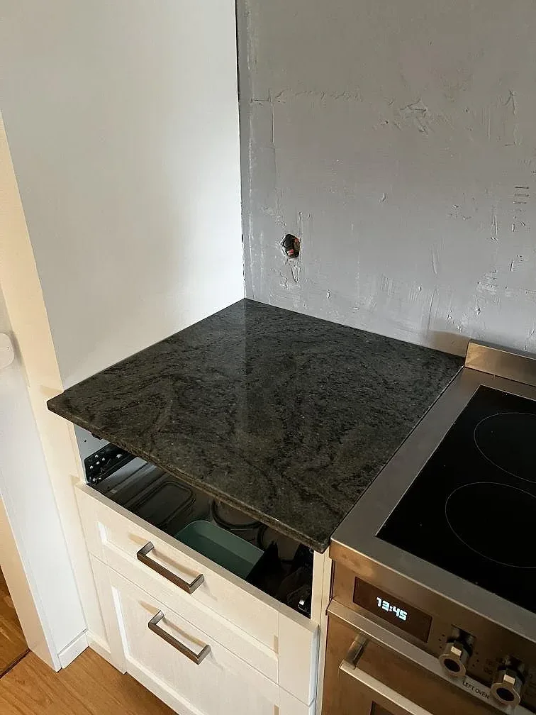 Kort bänksektion i polerad Verde Savana granit intill svart induktionshäll och vita shakerluckor i Limhamn.