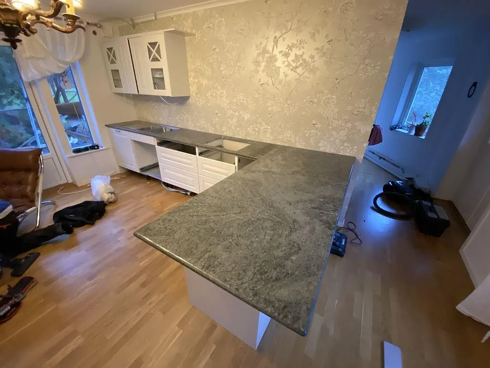 Köksbänkskiva i grön Verde Savana-granit, polerad, som löper runt hörnet till en stor halvö i ett kök med blommig tapet i Mariefred.
