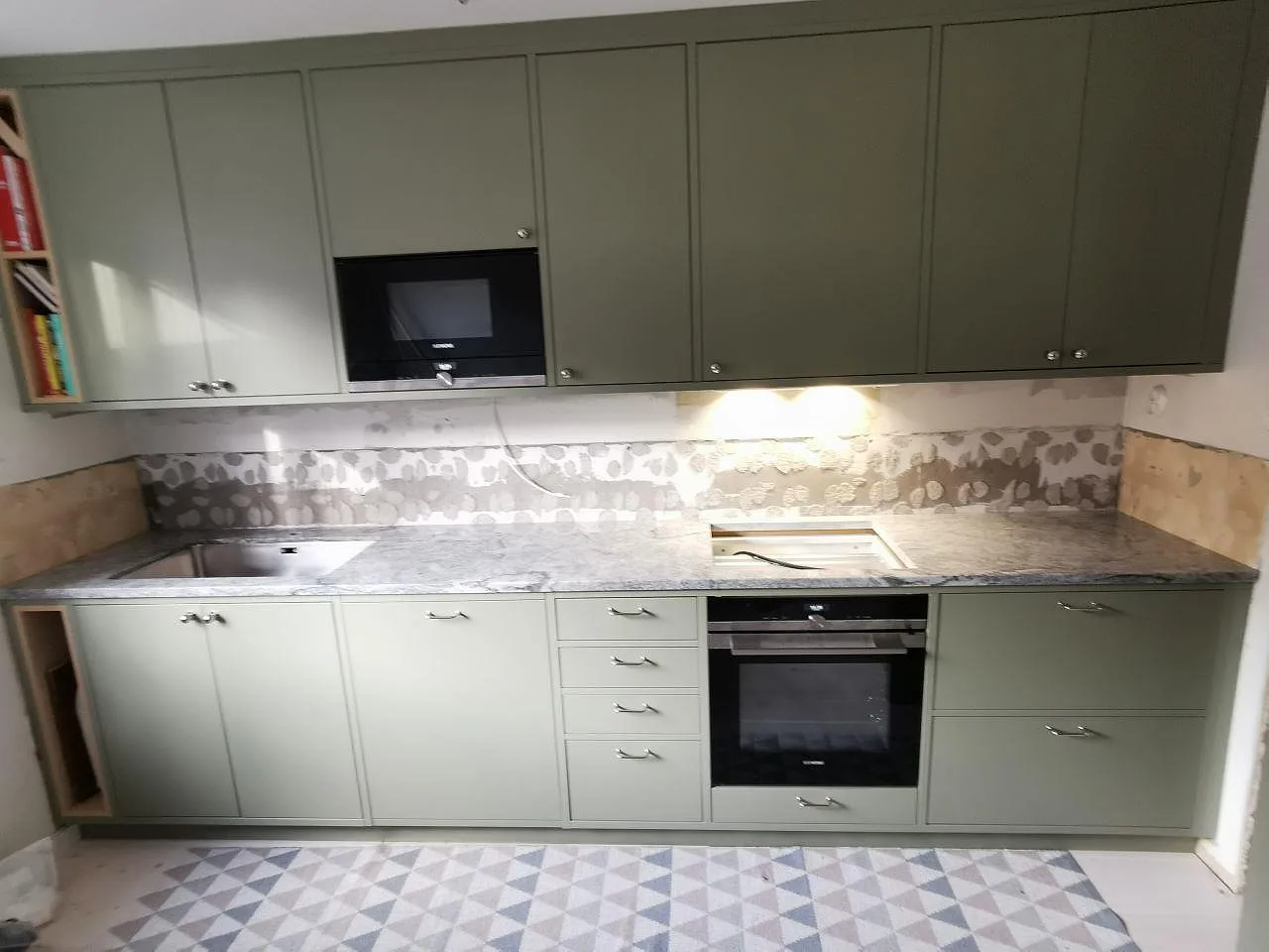 Köksbänkskiva i granit Viscont White, vit med grå ådring, nedsänkt diskho och mönstrat stänkskydd, mot salviegröna luckor i ett kök i Göteborg.