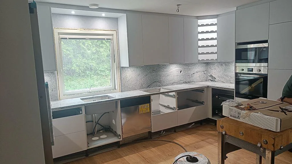Köksbänkskiva i Viscont White granit med grå-svart ådring, polerad, i ett L-format kök under montering i Hörby, med rostfri underlimmad diskho och matchande stänkskydd.