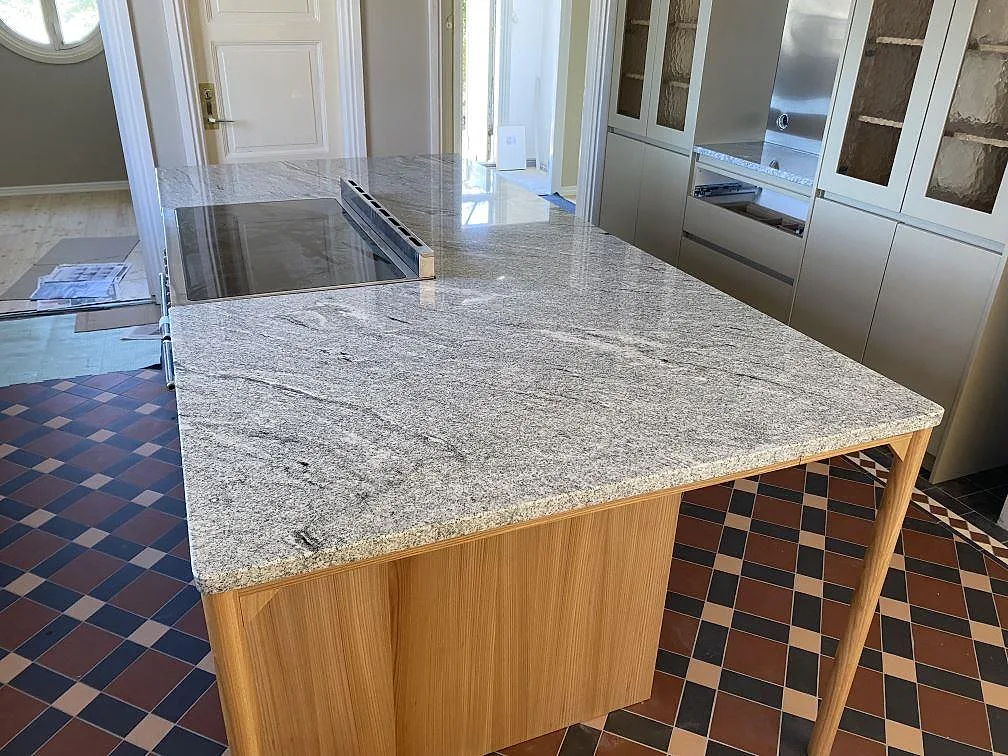 Köksö med polerad Viscont White granit, ljus köksbänkskiva med grå ådring och spis i ett kök under montering i Jämshög.