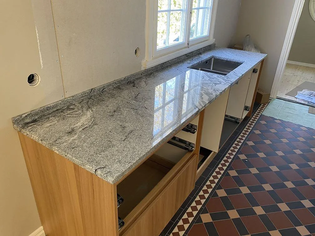 Lång köksbänkskiva i polerad Viscont White granit vid fönster, med diskho och öppen köksstomme i Jämshög.