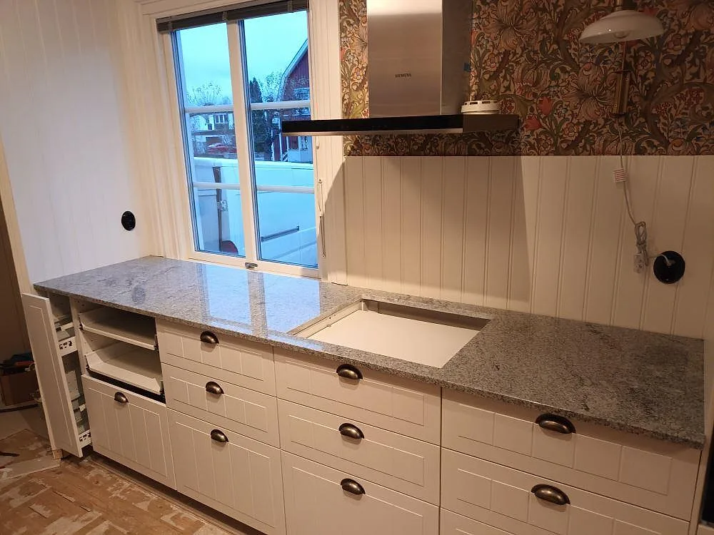 Köksbänkskiva i grå-vit Viscont White-granit, polerad, med plats för häll och uppvikt pärlspontvägg i ett kök under montering i Norrköping.