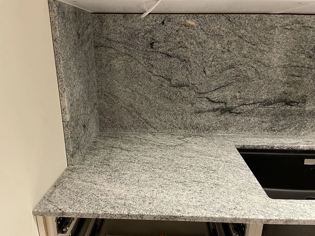 Hörndetalj av köksbänkskiva i Viscont White granit med ljusgrå ådring och uppvikt heltäckande stänkvägg i ett kök i Sollentuna.
