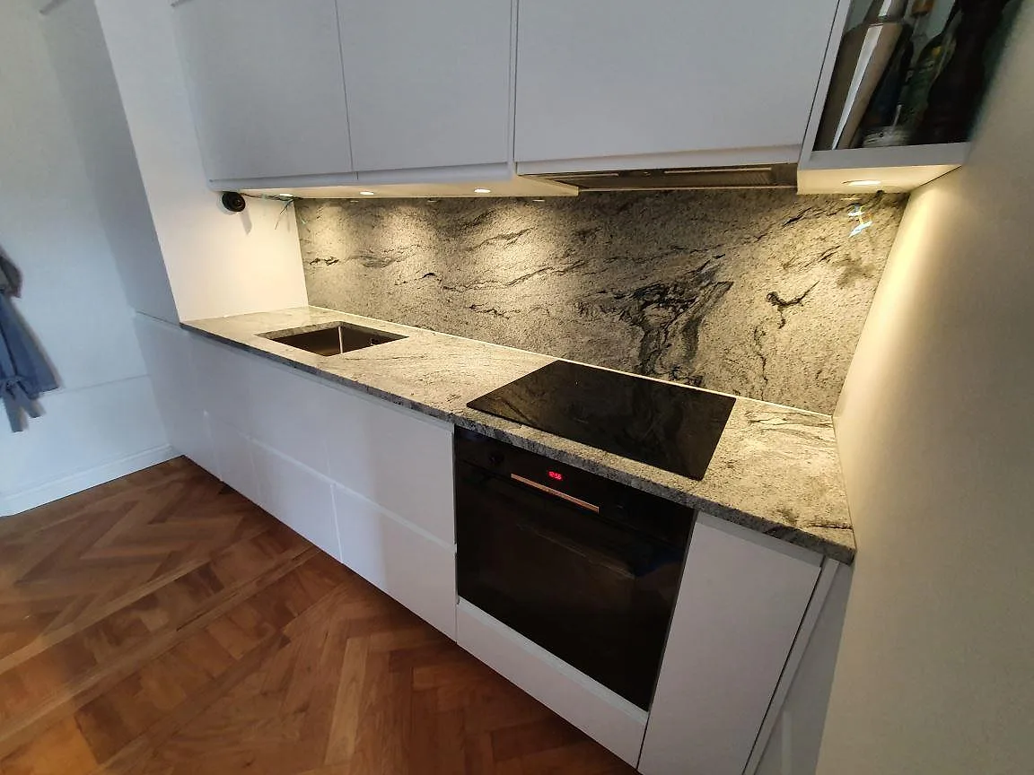 Enradigt kök i Stockholm med polerad Viscont White granitbänkskiva och matchande stänkvägg, vita greppfria luckor och fiskbensparkett, sett snett från sidan.