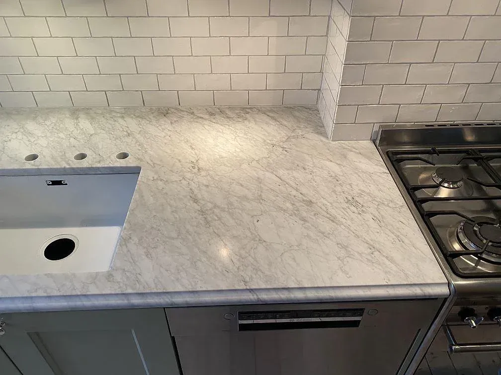 Köksbänkskiva i Carrara C marmor, matt, med underlimmad vit diskho och fristående rostfri gasspis i ett kök i Göteborg.