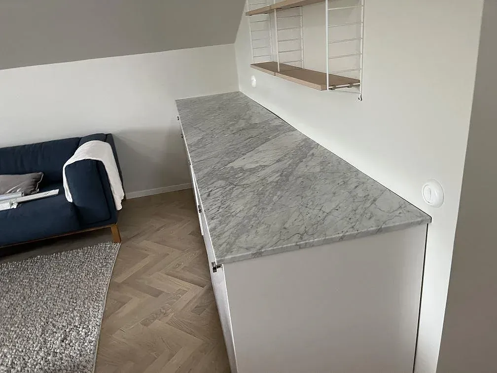 Bänkskiva i matt Carrara C med grå marmorådring, monterad ovanpå vita underskåp längs väggen i ett ljust rum under snedtak i Limhamn.