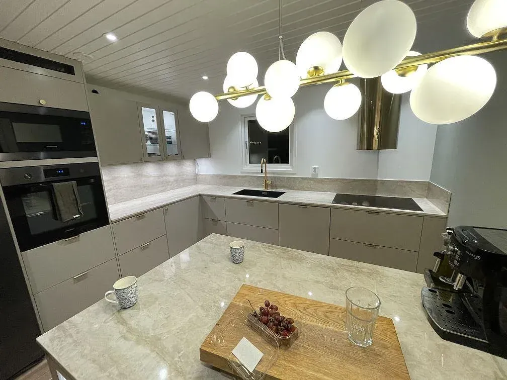Köksbänkskiva i beige Dekton Arga, polerad keramik med travertinlik ådring, i ett fininrett kök med köksö, svart underlimmad diskho, guldfärgad blandare och infälld induktionshäll i Billdal.