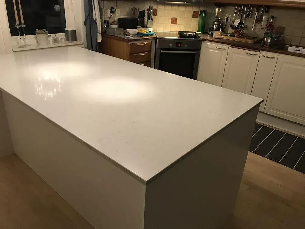 Köksbänkskiva i vit Silestone Lagoon, polerad kvartskomposit, på en fristående köksö med fristående spis och köksinredning i bakgrunden i ett kök i Pixbo.