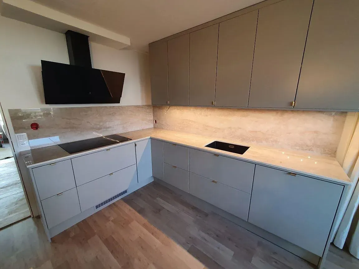 Köksbänkskiva i Dekton Arga, polerad beige keramik med travertinlik ådring, i ett L-format kök i Knutby med infälld induktionshäll, underlimmad diskho och heltäckande stänkskydd i samma sten.