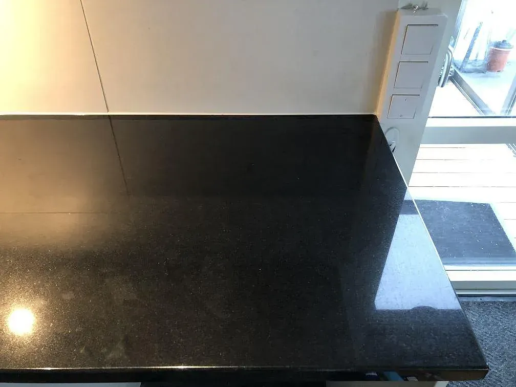 Köksbänkskiva i svart Nero Assoluto granit, polerad, som löper runt ett hörn vid fönstret i ett kök i Malmö.