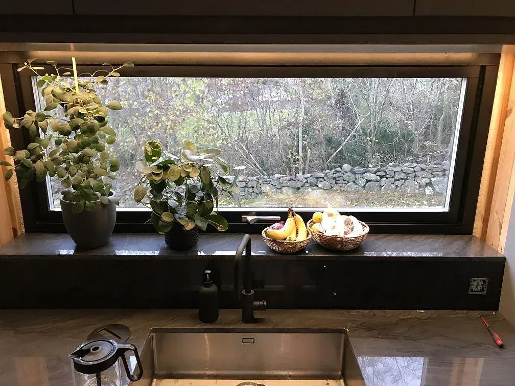 Köksbänkskiva i Sensa Nilo granit, polerad, med underlimmad rostfri diskho och matt svart blandare framför ett brett fönster mot trädgården i ett kök i Åsa.