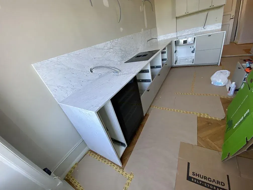 Köksbänkskiva i vit Carrara C marmor, matt, längs en hel köksvägg med infälld induktionshäll och uppvikt stänkkant under montering i ett kök i Malmö.